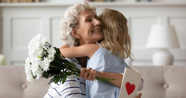 Hugs Grandmother and child hugging while child holds white flowers and a card with a red heart, expressing affection.